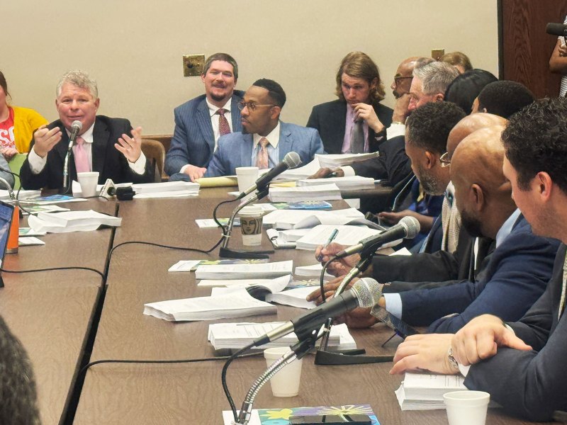 A group of people seated around a large, brown table. Placed on the table are microphones and several stacks of paper.