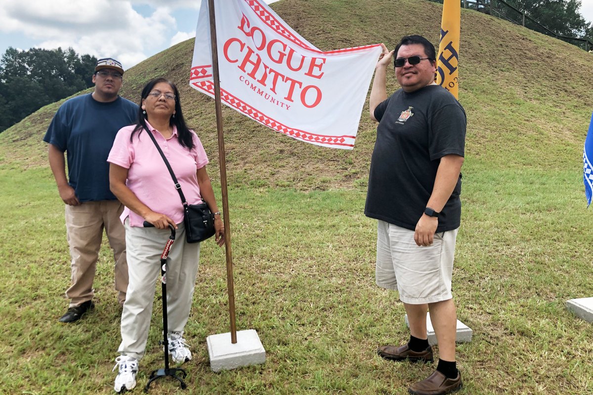 Three people stand by a mound with a flag that reads Bogue Chitto Community