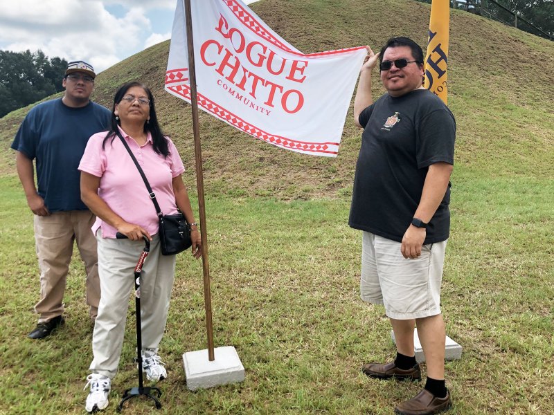 Three people stand by a mound with a flag that reads Bogue Chitto Community