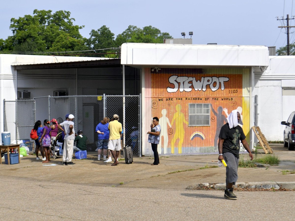 People walk around a building with a hand painted sign reading STEWPOT We are the Rainbow of the World