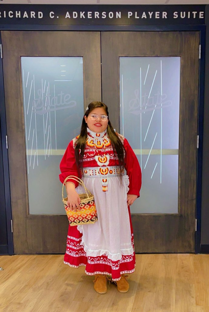 A young woman in a red and white dress of native design poses in front of a basketball player suite
