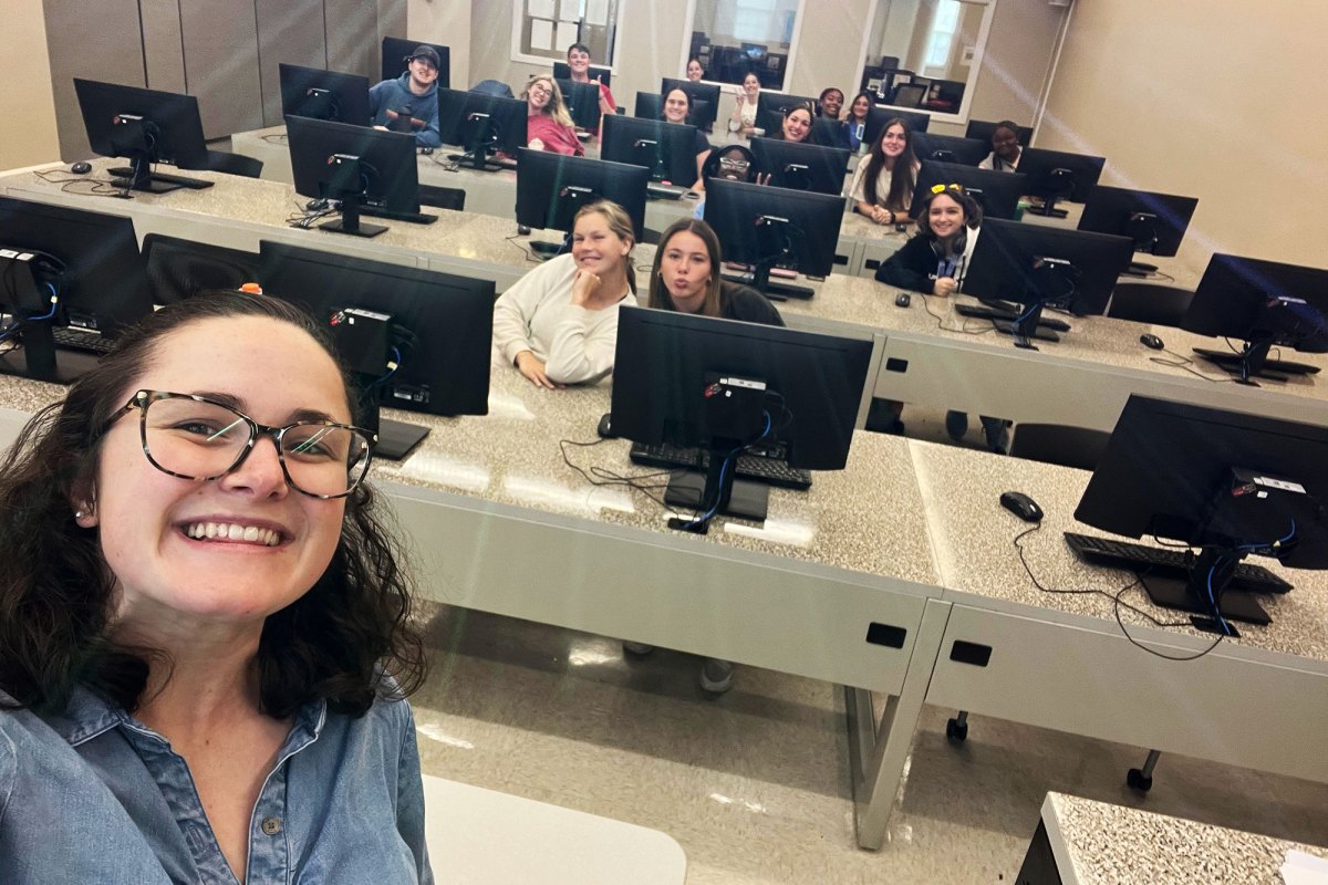 A woman takes a selfie at the front of a classroom full of students sitting at long tables covered in computers