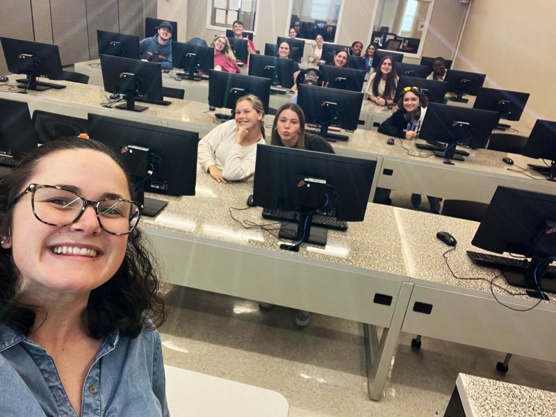 A woman takes a selfie at the front of a classroom full of students sitting at long tables covered in computers
