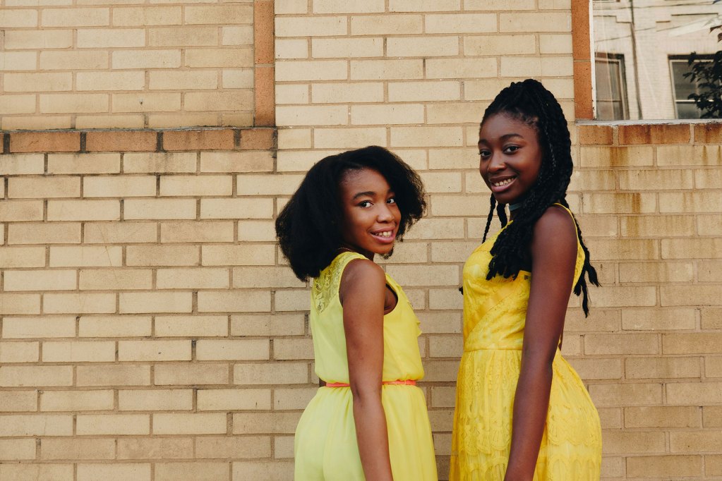 Two black girls in yellow dresses smile outside by a beige brick wall