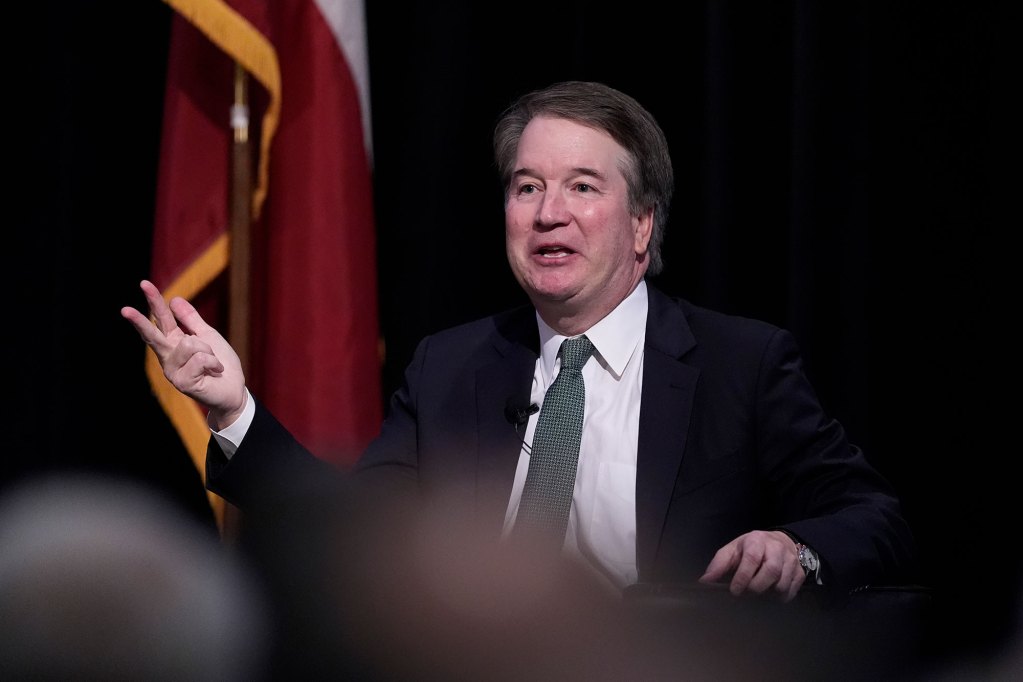 A man in a suit sits on a stage and talks. A US Flag can be seen standing to his right.