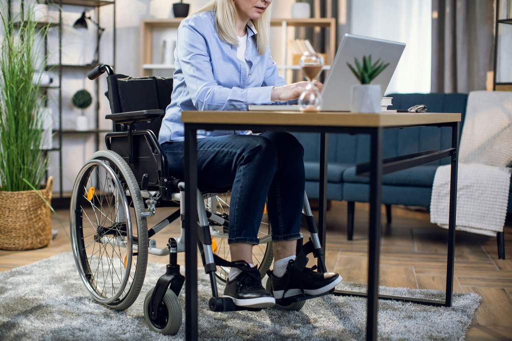 Woman wearing blue clothes sitting in a wheelchair, working on a laptop at a desk