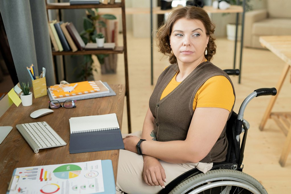 Young woman in orange and brown clothes, sitting in a wheelchair sitting at a work desk