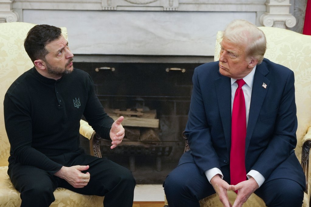 Two men sit in front of a white fireplace. The man in the left is in all black, the man on the right is in a suit and tie.
