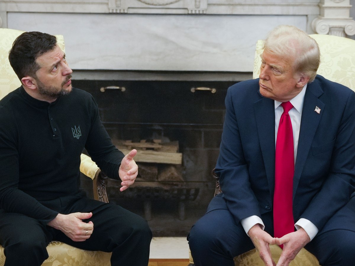 Two men sit in front of a white fireplace. The man in the left is in all black, the man on the right is in a suit and tie.
