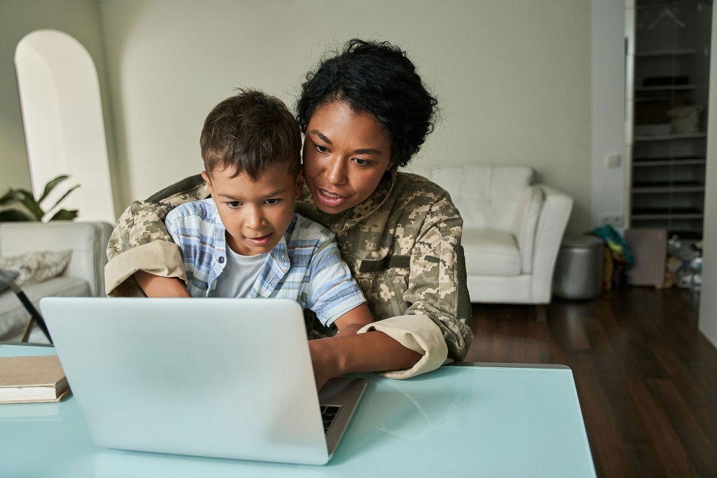 A young boy sits on the lap of a woman in military clothes, typing on a laptop