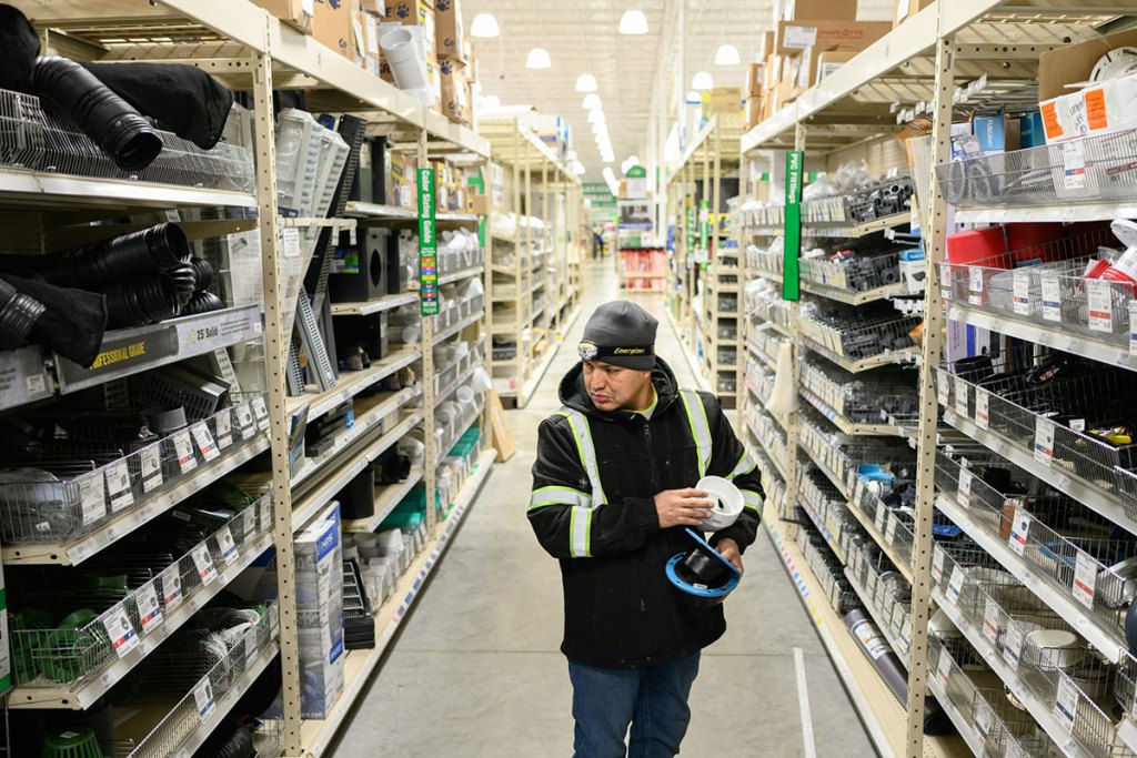 A man wearing a hat and a jacket holds items and walks down an empty aisle of what looks like a hardware store.