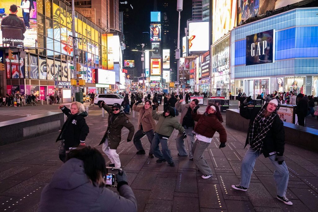 A group of people are seen moving on a busy New York City street, with many bright lights and billboards around them on the buildings