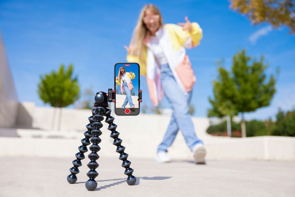 A smartphone on a mount on the ground shows an image of a young woman doing a dance move on a sunny day outside.