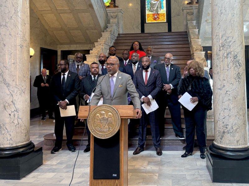 A man speaks at a podium bearing the seal of the State of Mississippi as several legislatures stand behind him and listen