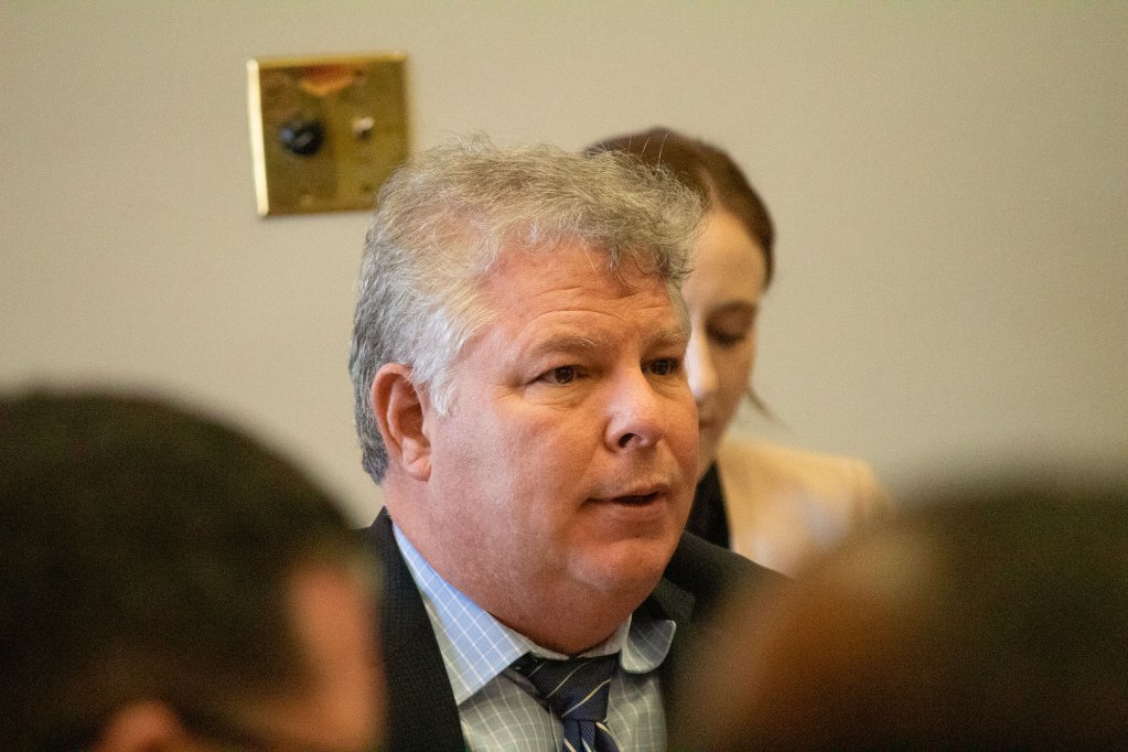 Man with white hair speaking in a room with two people in front of him and one woman behind him.