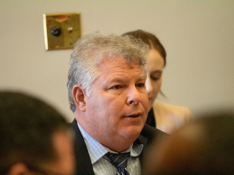 Man with white hair speaking in a room with two people in front of him and one woman behind him.
