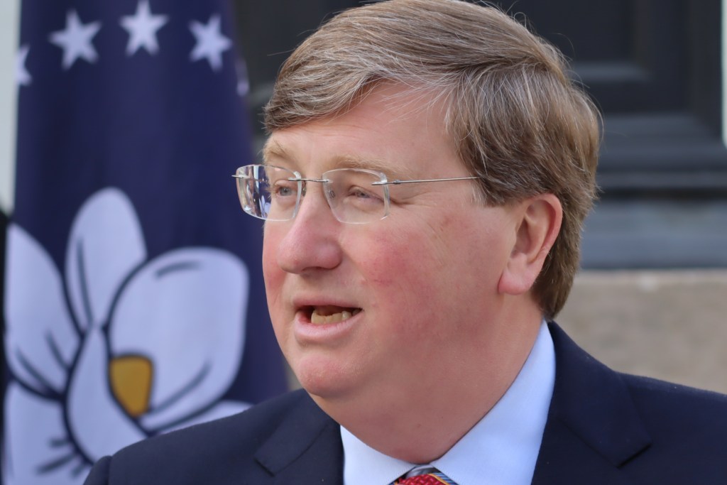 A photo of Tate Reeves speaking outside, a state of Mississippi flag visible behind him