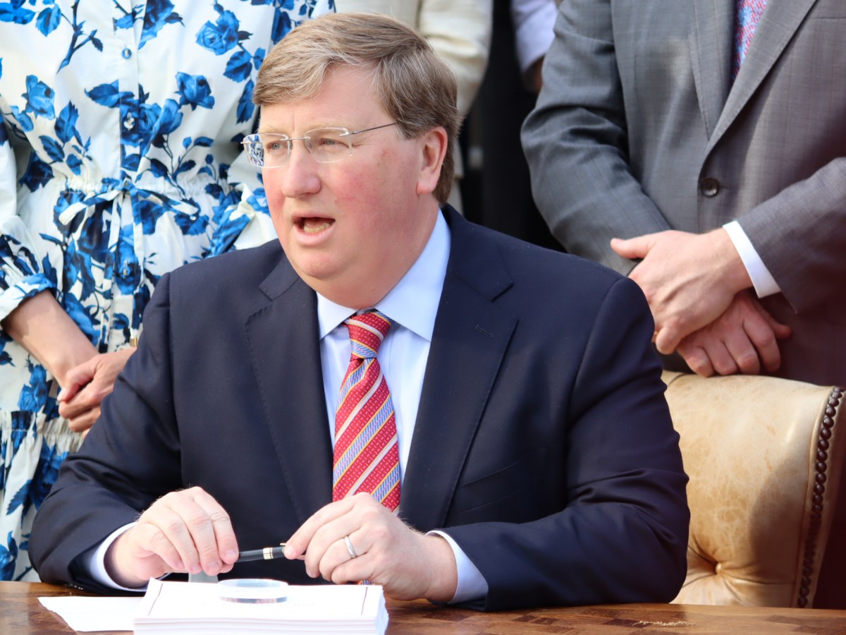 a photo of a man sitting at a table holding a pen in front of a stack of paper, with people standing behind him clasping their wrists with their opposite hands