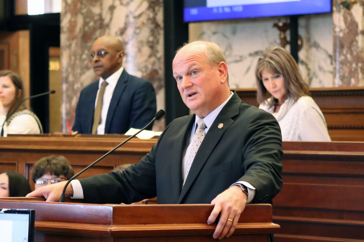 A man in a dark suit stands at a podium in the legislature, others visible at desks behind him as well