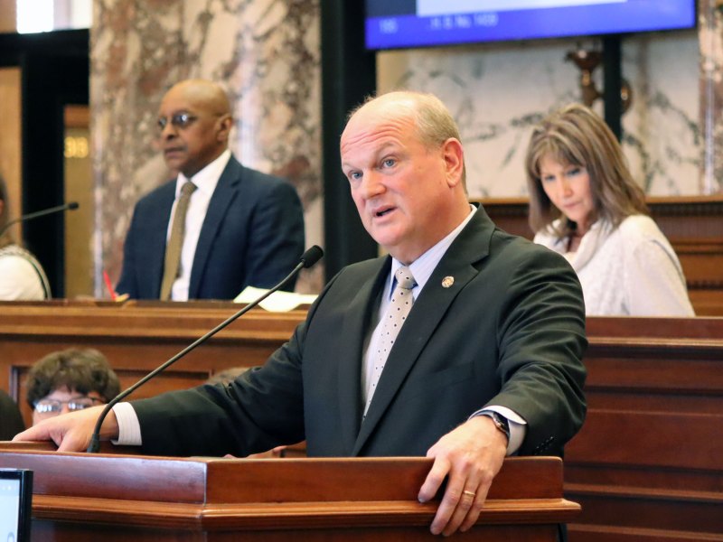A man in a dark suit stands at a podium in the legislature, others visible at desks behind him as well