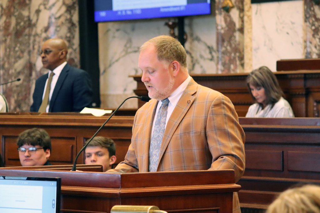 A man in a brown plaid suit stands at a podium speaking into a microphone.