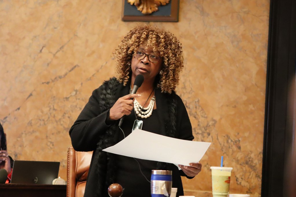 A woman holding a mic wearing black with pearls around her neck