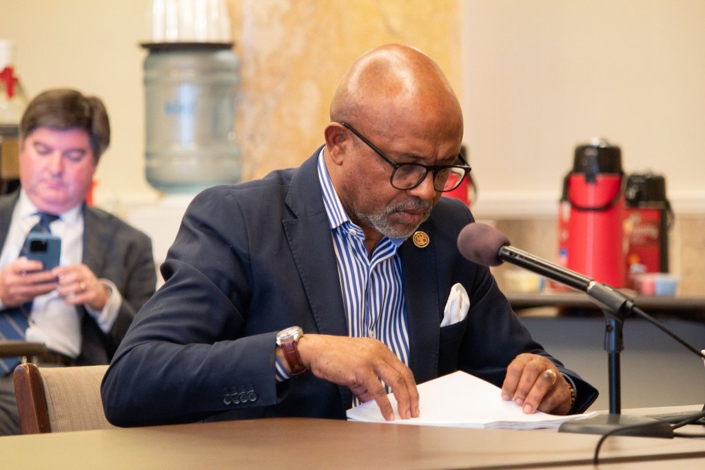 House Representative Robert Johnson speaks at a mic while wearing a navy suit jacket and a blue/white shirt.