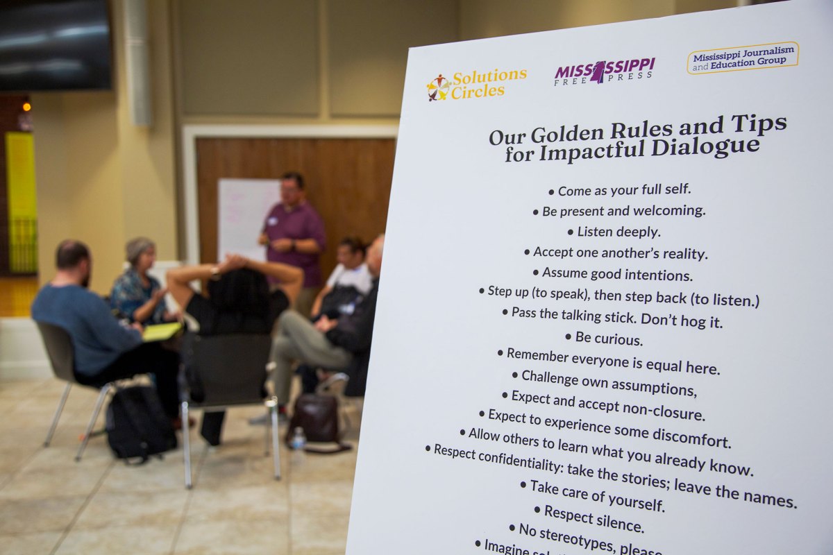 Mississippi Free Press poster pictured in front right with people sitting at a table behind it.