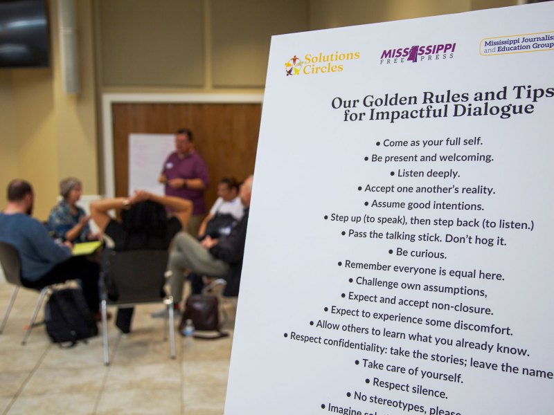 Mississippi Free Press poster pictured in front right with people sitting at a table behind it.