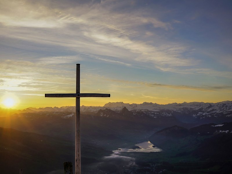 A wooden cross stands tall with sun and sky behind it.