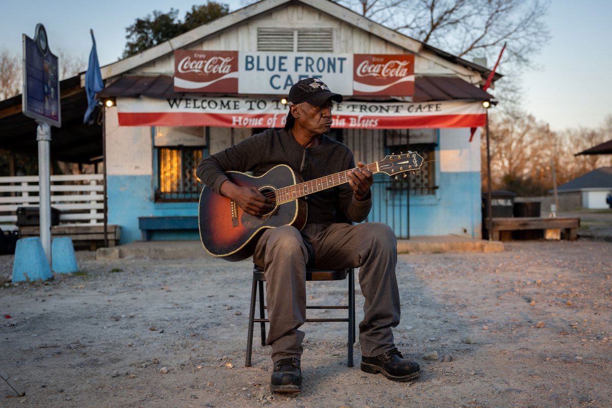 Jimmy Duck Holmes wearing a black hat and shirt plays a guitar while sitting in front a white and blue building.