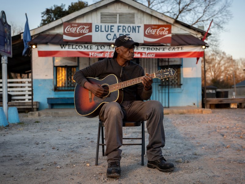 Jimmy Duck Holmes wearing a black hat and shirt plays a guitar while sitting in front a white and blue building.