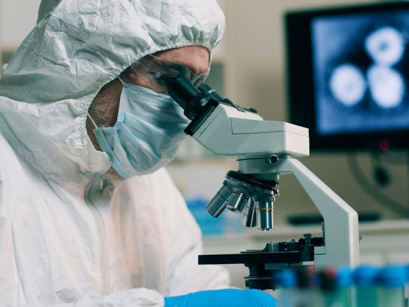 A person in full lab scrubs and head covering looks into a microscope
