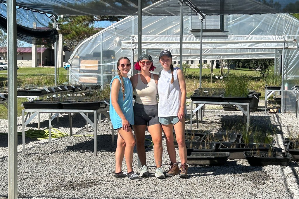 Three people stand outside of a plant nursery on a sunny day