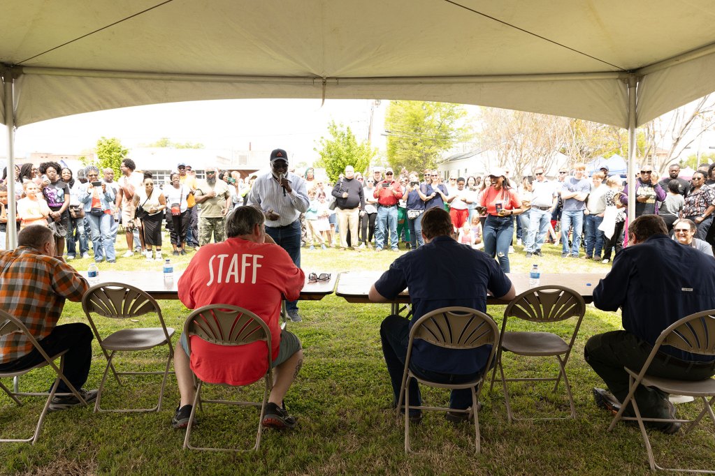 Men sitting at a table under a tent eating food while people outside of the tent in front of them watch