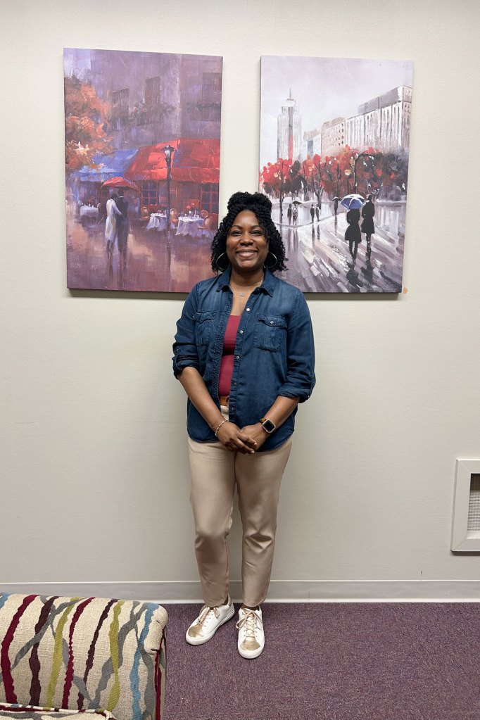 A woman in a blue jean shirt stands in front of two paintings on a wall.