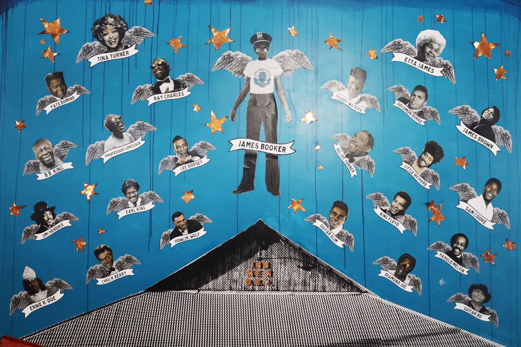 A blue wall pictured with a mix of blues, rock and roll, and R&B legends whom passed away.