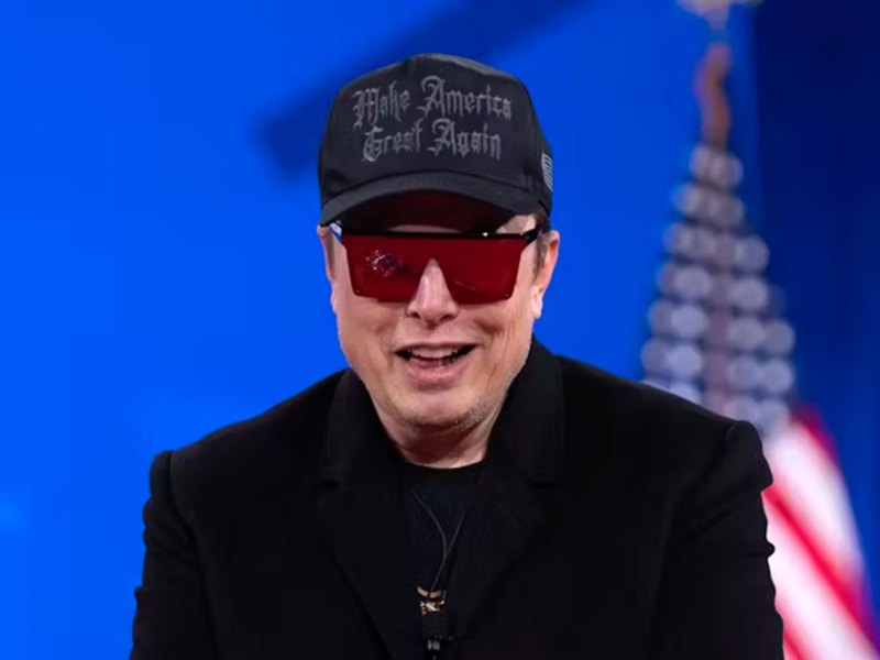 Elon Musk wearing a black hat and red glasses sits in front of blue wall and American flag.