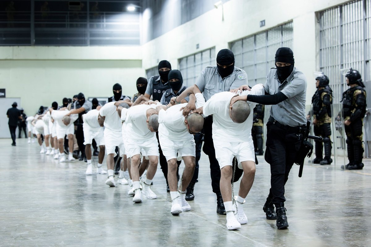 Masked guards transfer people all dressed in identical white shirts and shorts with chains around their legs and hands, heads shaved bald.