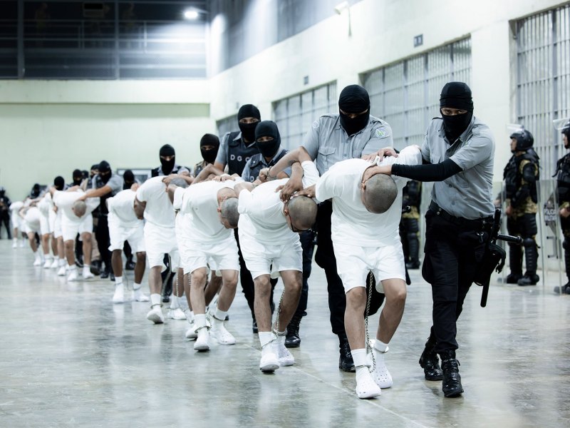 Masked guards transfer people all dressed in identical white shirts and shorts with chains around their legs and hands, heads shaved bald.