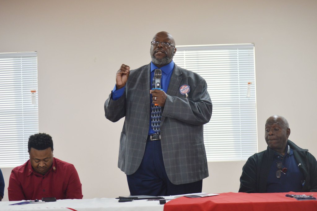 A man in a grey suit jacket and blue shirt speaking while two men sit to his left and right.