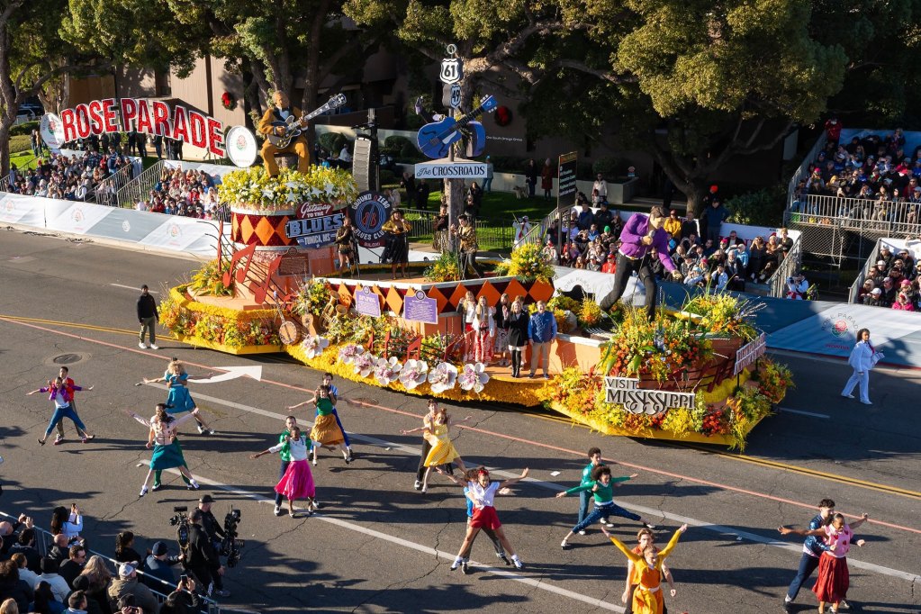 Men and women in colorful clothes dance on a street in front of a yellow float.