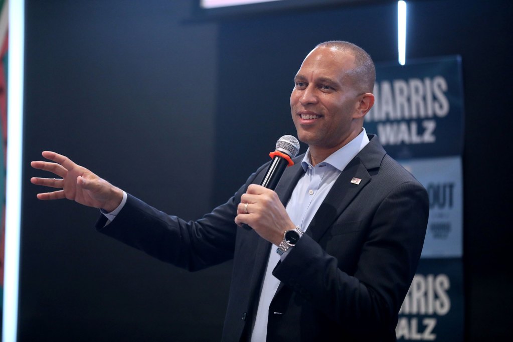 Hakeem Jeffries speaking with a mic in hand wearing a navy suit jacket and blue shirt.