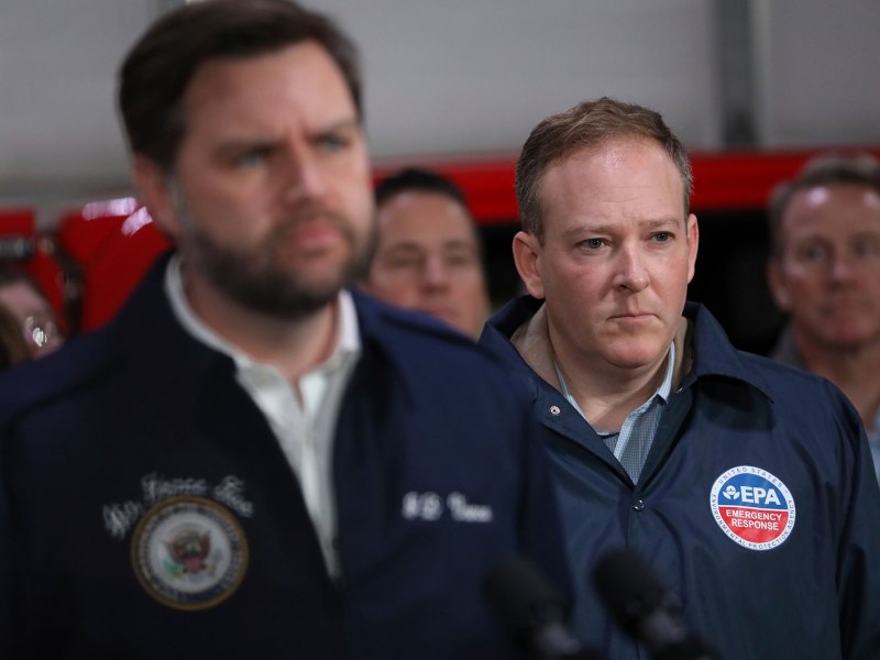 An indoor scene of me in blue jackets listening as one speaks at a press podium