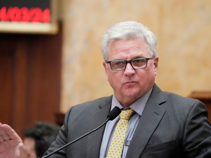 A man in grey suit and yellow tie speaks at a mic with one hand raised out