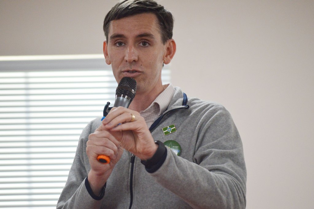 A man in a grey sweatshirt holds a grey mic while speaking