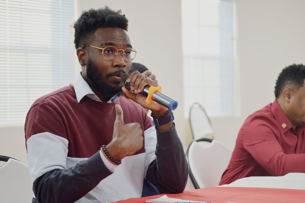 A man with a striped shirt and glasses speaking while holding a grey mic.