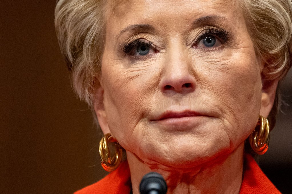 A closeup of Linda McMahon at a mic, wearing a red top and gold earrings