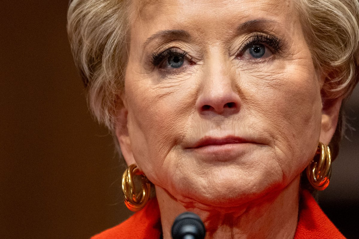 A closeup of Linda McMahon at a mic, wearing a red top and gold earrings