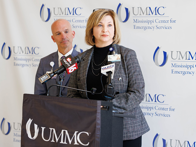 A woman and a man stand at a podium labeled UMMC with news mics clipped to the podium